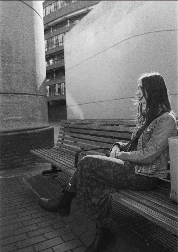 This picture is in black and white, shows a woman smartly dressed sitting on a bench with a tall bag near her. Her face is partially covered with her hair and we can see her profile. There is a Brutalist style concrete wall behind her and a pillar on one side.